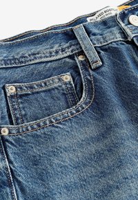 Gros plan d’un jean en denim bleu montrant la poche avant, la passant de ceinture, les coutures, les rivets métalliques, le bouton et l’étiquette portant la mention "Relaxed Bootcut Mid Rise."