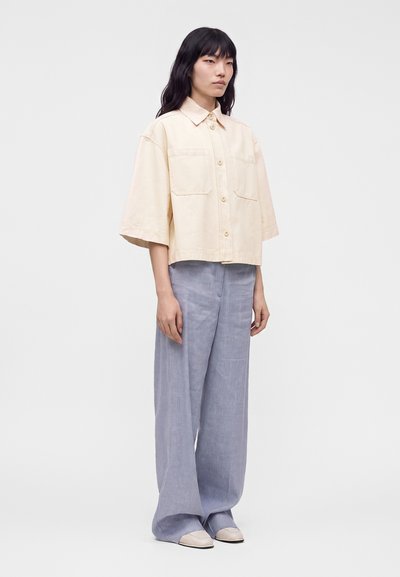 Femme aux cheveux noirs portant une chemise beige ample à manches courtes boutonnée et un pantalon large bleu clair, debout devant un fond uni.