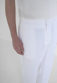 Bijele lanene hlače s glatkom teksturom, naborima i zatvaranjem na zip. Prikaz izbliza s rukom koja se oslanja na tkaninu.