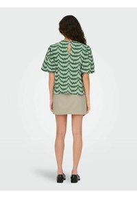 Blouse à manches courtes verte à motif avec un détail dos en trou de serrure, associée à une mini-jupe beige. Le matériau semble léger et tissé.