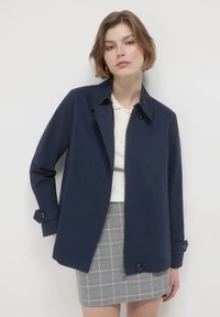 Giacca blu navy realizzata in tessuto liscio, con colletto, zip frontale e dettagli in bottone sulle maniche. Abbinata a una gonna a quadri.