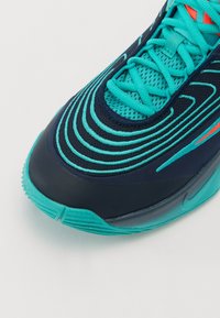 Gros plan d'une chaussure de sport bleu sarcelle et bleu marine avec des motifs cousus ondulés et des lacets bleu sarcelle sur fond blanc.