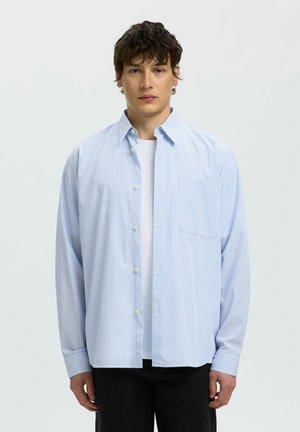 Jeune homme aux cheveux courts, bouclés et foncés, portant une chemise à rayures bleu clair boutonnée par-dessus un t-shirt blanc et un pantalon noir, sur fond blanc.