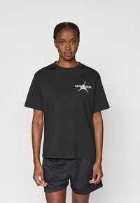 Μαύρο βαμβακερό T-shirt με χαλαρή εφαρμογή, που διαθέτει ένα χρυσό και λευκό λογότυπο "GENERATION" και σιλουέτα στην αριστερή περιοχή του στήθους.