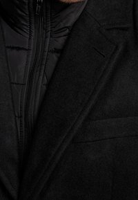 Gros plan sur un manteau en laine noire superposé à une veste matelassée noire avec une fermeture éclair centrale, montrant les détails du col et du revers.