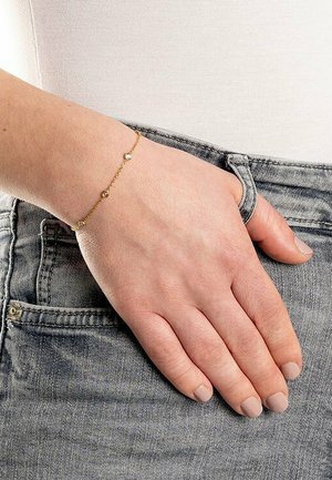 Bracciale in oro con piccoli perline rotonde distribuite uniformemente lungo la catena. Mano appoggiata su un denim grigio con una maglietta bianca visibile.