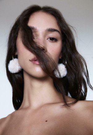 Modella con lunghi capelli scuri che parzialmente coprono il viso, indossa grandi orecchini con pompon bianchi e soffici su cerchi dorati. Pelle liscia, trucco naturale.