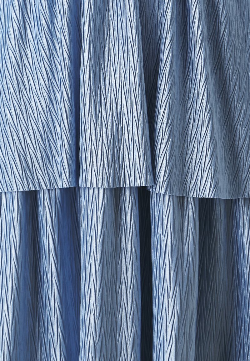 Jasnoniebieska, teksturowana tkanina o falistym wzorze, z ukośnymi liniami i warstwami. Miękka w dotyku, z płynącym wyglądem.