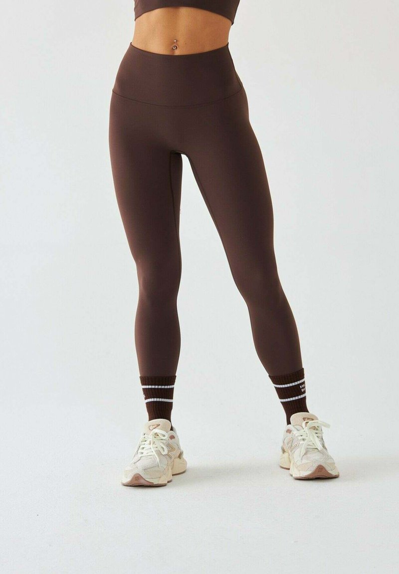Brązowe legginsy z wysokim stanem o gładkiej fakturze, zestawione z białymi i brązowymi butami sportowymi oraz brązowymi skarpetkami w paski.