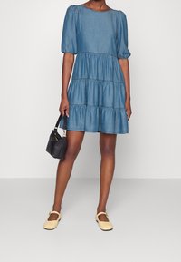 Vestido de mezclilla con mangas cortas abullonadas, diseño de volantes en capas y escote redondo. Lleva un bolso negro y zapatos planos de color amarillo claro.