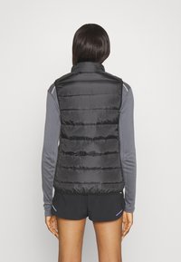 Juodas pūkuotas liemenė su aukštu apykakle, siuvinėtu dizainu ir glotnia apdaila, dėvima ant pilkos ilgomis rankovėmis sumuštinio, demonstruojanti prigludusią siluetą.