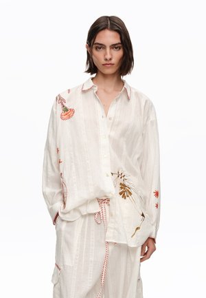 Joven con camisa blanca suelta abotonada con bordado floral y pantalones blancos a juego con cordón rojo estampado.