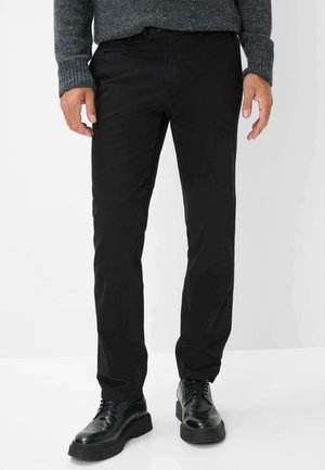 Schwarze Hose mit geradem Schnitt, ausgestattet mit Seitentaschen und einem Bund mit Gürtelschlaufen. Glatte Stofftextur, ergänzt durch schwarze Lederschuhe.