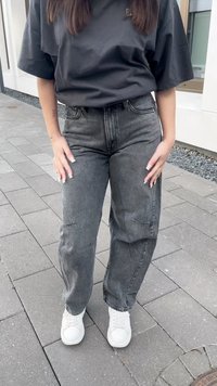 Grå høytlivsjeans med løs passform og små slitasjer; paret med hvite sneakers. Bakgrunn av grå brosteinsheller.