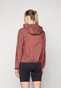 Vandafvisende, cropped jakke i dyb burgunder med hætte, elastiske manchetter og en subtil glans. Kombineret med sorte cykelshorts.