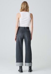 Jeans neri a gamba larga con risvolti e vita media, abbinati a una canotta bianca. Il tessuto è denim con una texture liscia.