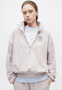 Felpa rosa con zip e colletto alto, con un patch rotondo che dice "PARIGI". Indossata sopra una camicia azzurro chiaro, texture morbida.