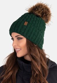 Gorro de punto verde con un patrón texturizado, que presenta un gran pompón de piel sintética en la parte superior. Detalles adicionales incluyen una etiqueta de cuero marrón.
