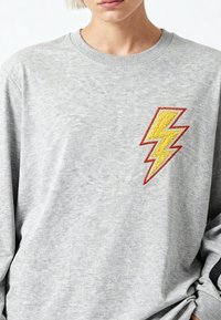 Person trägt hellgrauen Pullover mit gelbem Blitz-Aufnäher, rot umrandet, auf der Brust vor einfachem hellem Hintergrund.