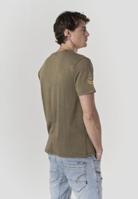 T-shirt di cotone verde oliva con scollo arrotondato, maniche corte e toppa decorativa sulla manica sinistra. Vestibilità casual e tessuto liscio.