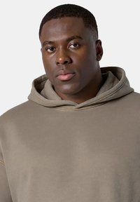 Jasnobrązowy hoodie z kieszenią typu kangur, o miękkiej fakturze i kapturem ze sznurkiem. Charakteryzuje się luźnym krojem i widocznymi akcentami szwu.