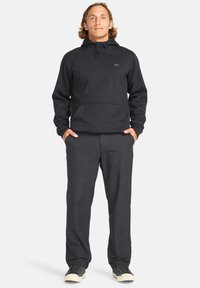Felpa nera con tasca frontale, cappuccio con coulisse e patch con logo. Abbinata a pantaloni neri coordinati. Tessuto texturizzato e vestibilità rilassata.