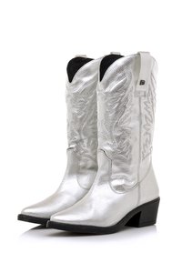 Botas de vaquero plateadas con puntas puntiagudas, hechas de cuero brillante. Presentan diseños de costuras elevadas y parte superior curva con tiradores. Tacones bajos de bloque.