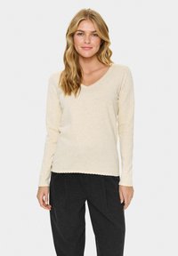 Gai beidzama bezgaļas džemperis ar V-veida izgriezumu un viļņu apmalēm. Mīksta tekstūra, brīva piegriezuma stils, valkāts ar tumšām biksēm. Nav izteiktu rakstu vai akcentu.