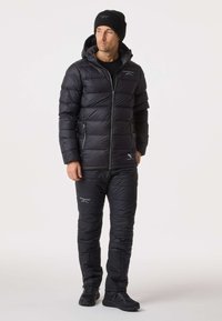 Svart puffjacka med huva, dragkedja framtill och quiltat mönster, tillsammans med matchande quiltade byxor och svarta skor.