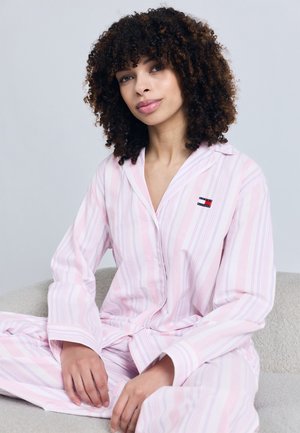 Jauna moteris su garbanotais plaukais sėdi, dėvi šviesiai rožinės ir baltos spalvos dryžuotas naktinukes su mažu logotipu krūtinėje.