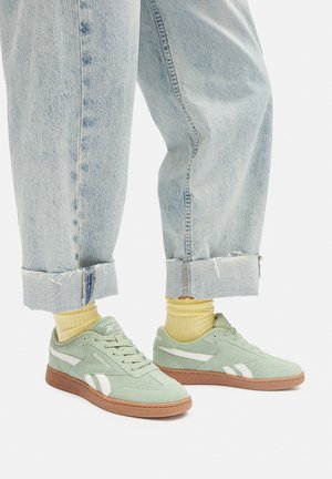Jasnozielone trampki z białymi akcentami, wykonane ze skóry zamszowej i siateczki, z okrągłym noskiem, płaską brązową gumową podeszwą, stylizowane z podwiniętymi jasnoniebieskimi dżinsami i żółtymi skarpetkami.