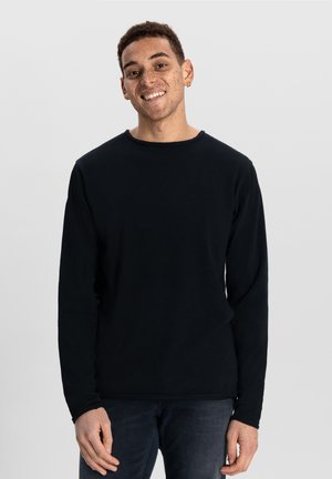 Tricou negru cu mânecă lungă din material moale, cu guler rotund și croială lejeră, purtat cu blugi negri.