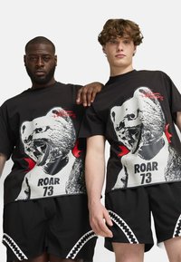 Čierne tričká obsahujú grafiku búrlivého medveďa v bielej a červenej farbe s textom "ROAR 73." Sú skombinované s čiernymi šortkami z oka s bielymi akcentmi.