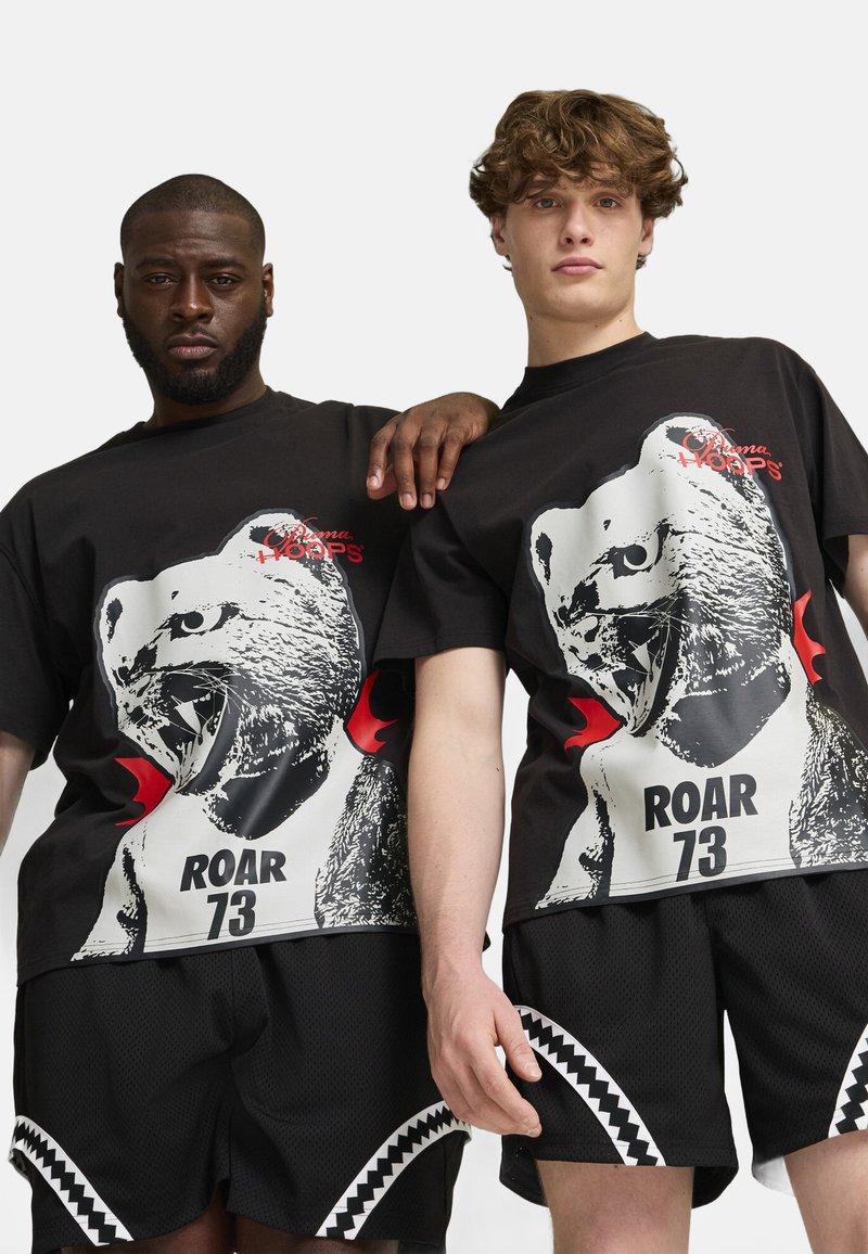 Čierne tričká obsahujú grafiku búrlivého medveďa v bielej a červenej farbe s textom "ROAR 73." Sú skombinované s čiernymi šortkami z oka s bielymi akcentmi.