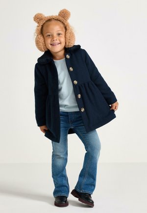 Bambino sorridente con cuffie in peluche a forma di orso beige, cappotto blu scuro con bottoni, camicia azzurra chiara, jeans blu e stivali neri, in piedi su uno sfondo bianco.