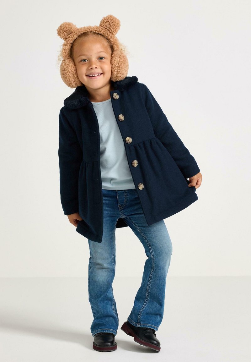 Bambino sorridente con cuffie in peluche a forma di orso beige, cappotto blu scuro con bottoni, camicia azzurra chiara, jeans blu e stivali neri, in piedi su uno sfondo bianco.