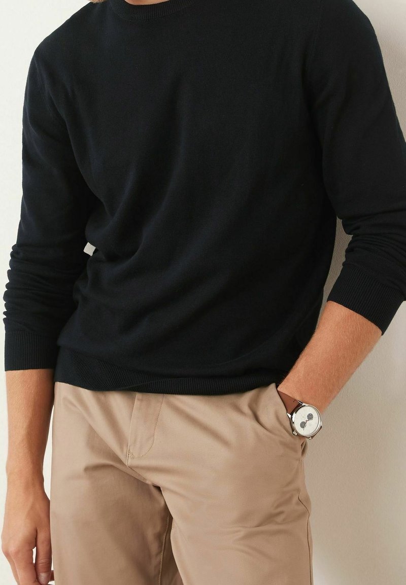 Homme portant un pull noir à manches longues et un pantalon beige, une main dans la poche, montrant une montre-bracelet argentée au poignet gauche.