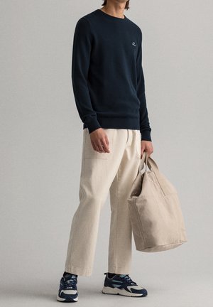 Man wearing navy sweater, beige wide-leg pants, and navy sneakers, holding a beige canvas tote bag against a plain gray background.