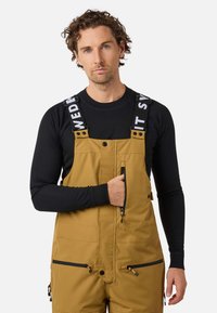 Mężczyzna z kręconymi włosami, ubrany w musztardowe ogrodniczki z czarnymi kieszeniami na zamek oraz czarną koszulę z długim rękawem, z ręką w kieszeni na piersi, na neutralnym tle.