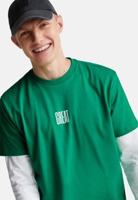 Camiseta de algodón verde de manga corta, con el texto "GREAT" en blanco. Usada sobre una camiseta de manga larga blanca y combinada con una gorra oscura.