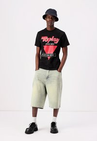 Camiseta de algodón negra con gráfico rojo y blanco de "Replay", combinada con pantalones cortos de mezclilla verde claro y zapatos negros gruesos.