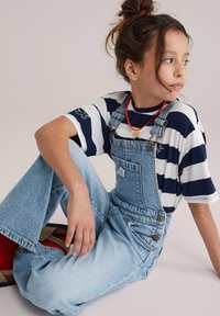 Salopetă din denim albastru deschis, cu bretele reglabile și buzunare laterale. Asortată cu un tricou cu dungi albastre și albe. Șosete roșii ca accente vizibile.