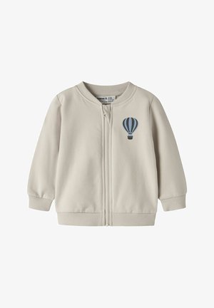 Veste zippée beige clair en coton, avec un col rond, des poignets côtelés, et un motif de montgolfière bleue sur le côté gauche.