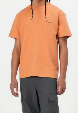 Osoba ubrana w pomarańczowy T-shirt Carhartt i ciemnoszare spodnie cargo, stojąca z dwoma opadającymi warkoczami.