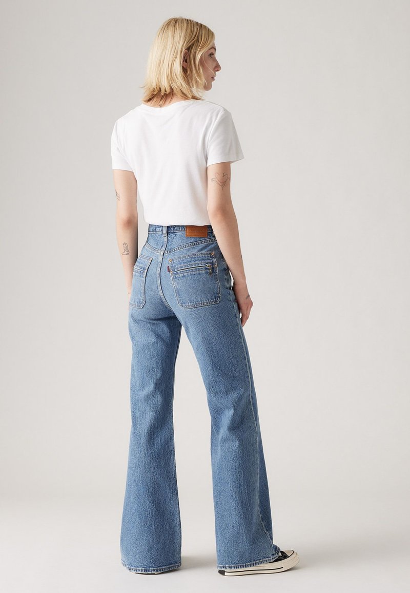 Jean en denim bleu clair taille haute évasée, avec des poches arrière et une patch en cuir marron à la taille. Porté avec un t-shirt blanc.