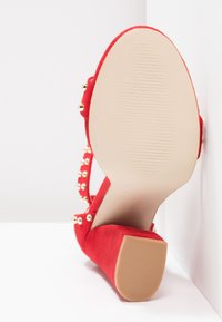 Zapato de tacón alto de ante rojo con punta redondeada, acentos de tachuelas en las correas del tacón y una suela de goma beige texturizada.