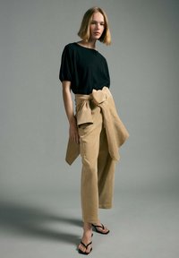 Woman with short blonde hair wearing a black top, beige pants, and a matching beige jacket tied around her waist, standing on a gray background.