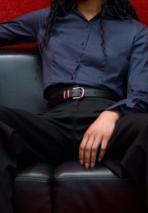 Person, der bærer en marineblå skjorte med knapper og sorte bukser med sort bælte, siddende på en sort lædersofa mod en rød baggrund.