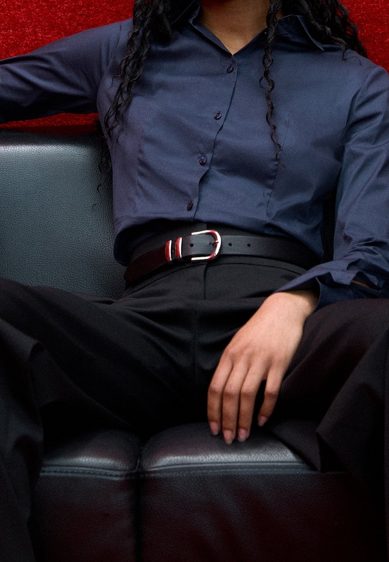 Personne portant une chemise bleu marine boutonnée et un pantalon noir avec une ceinture noire, assise sur un canapé en cuir noir sur fond rouge.