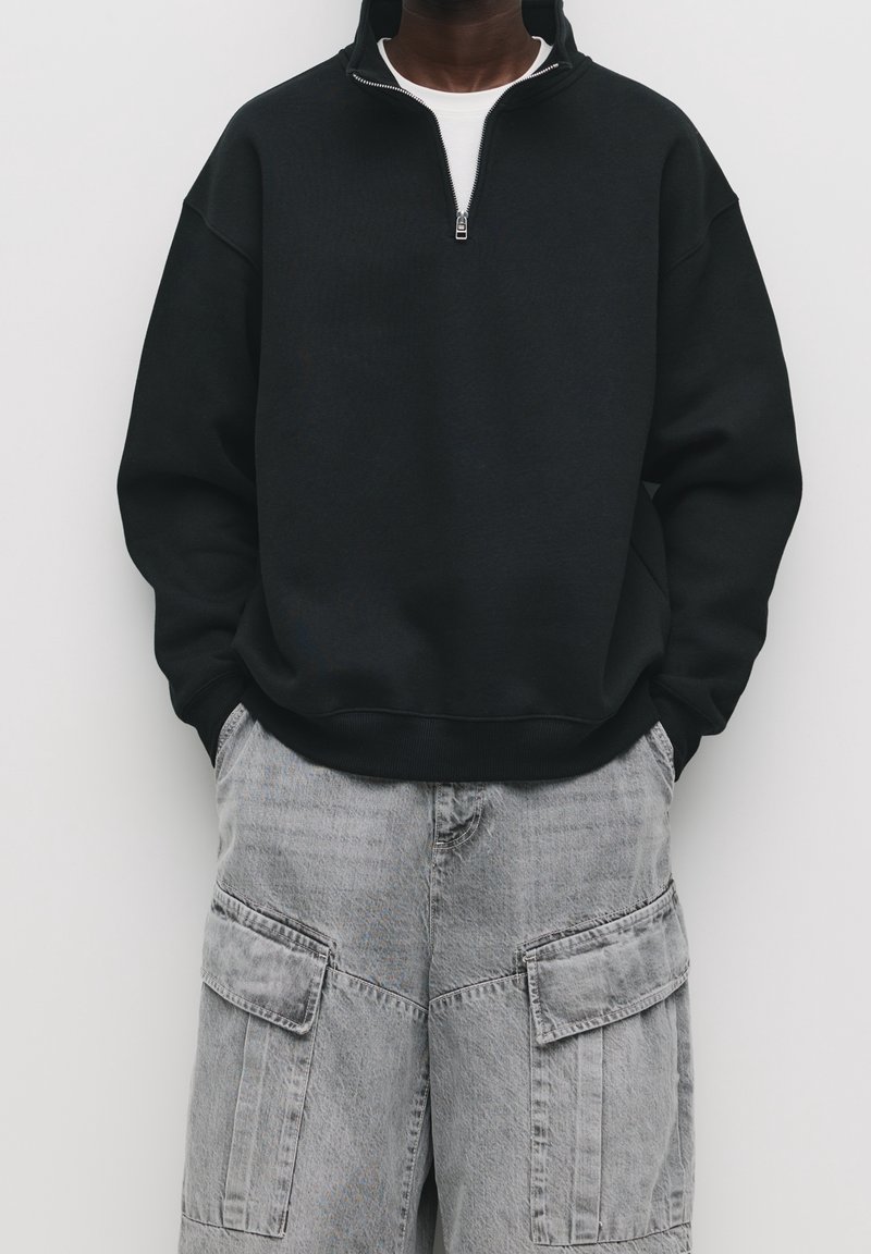 Personne portant un sweat-shirt noir à fermeture éclair quart, les mains dans les poches d'un pantalon cargo gris clair sur un fond uni.